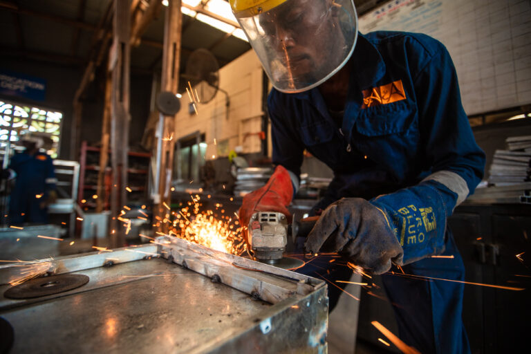 Fabricator in a face shield working a steel piece at the Accents & Art workshop, sparks across the bench